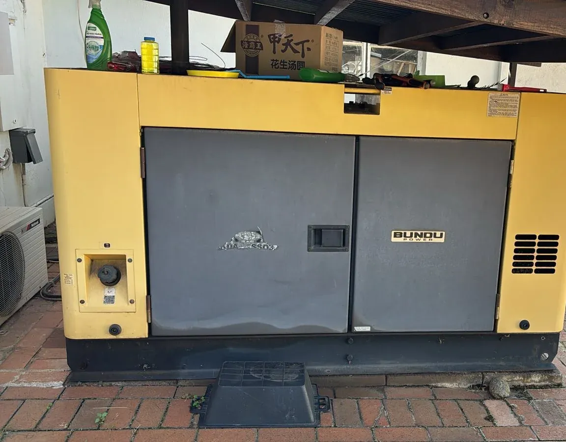 Yellow Bundu Power canopied diesel generator under a carport on brick paving, tools and bottles on the roof