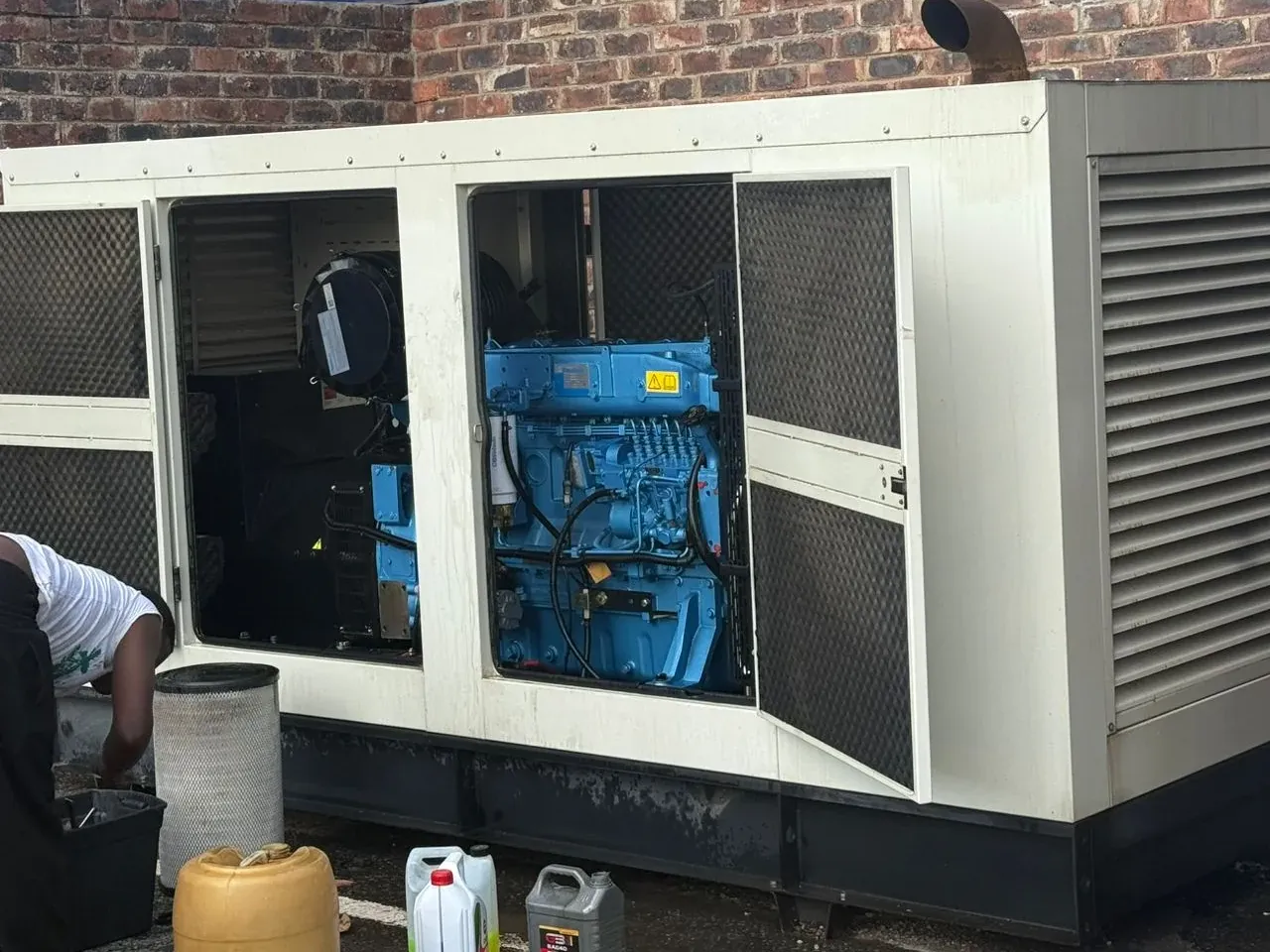Large cream canopied diesel generator with service doors open on a blue engine, one person working beside oil containers