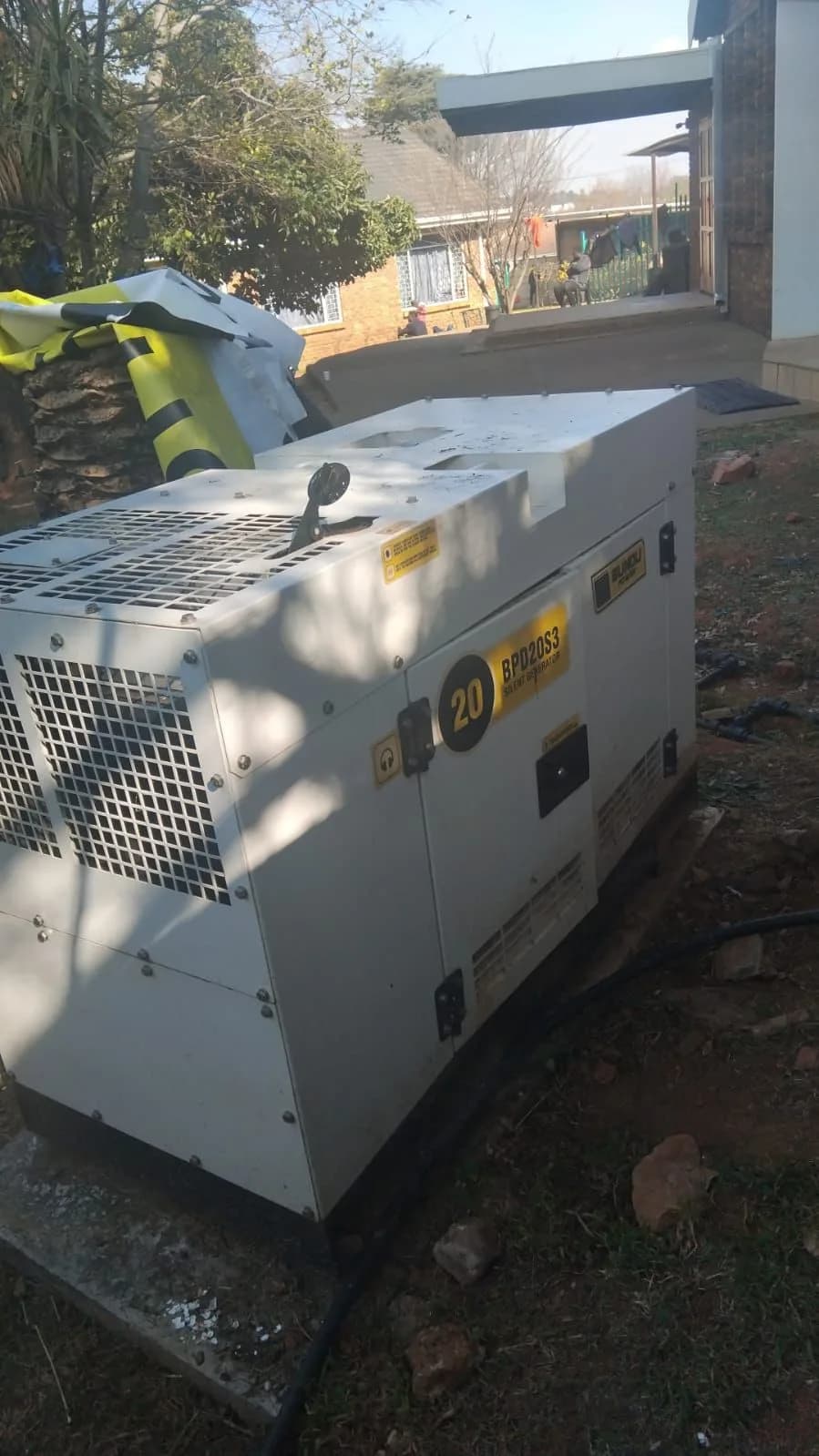 White Bundu Power 20 kVA silent diesel generator on bare ground beside a brick building, four people far behind