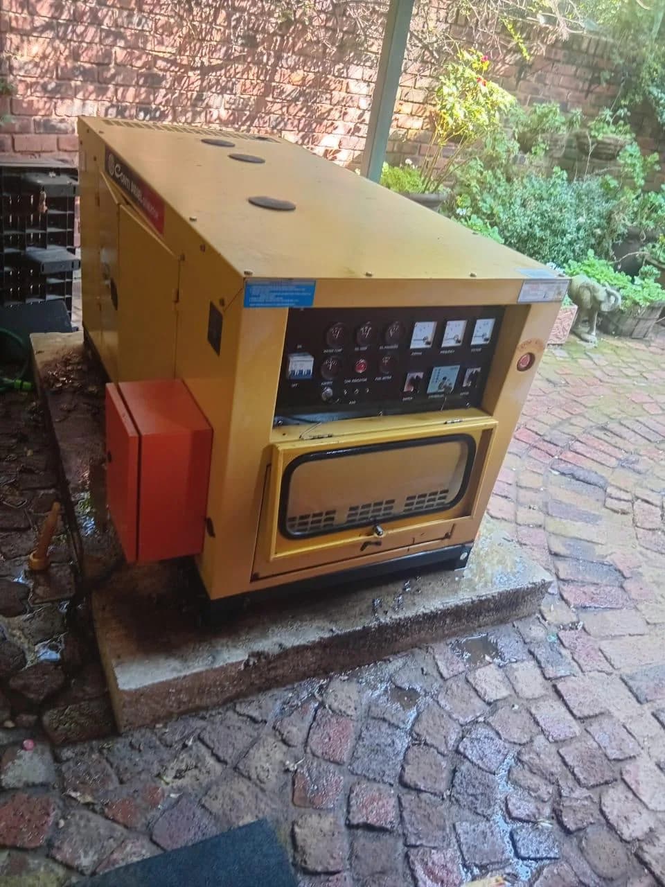 Yellow canopied diesel generator with analogue gauges and a red side box on a concrete plinth in a wet paved courtyard