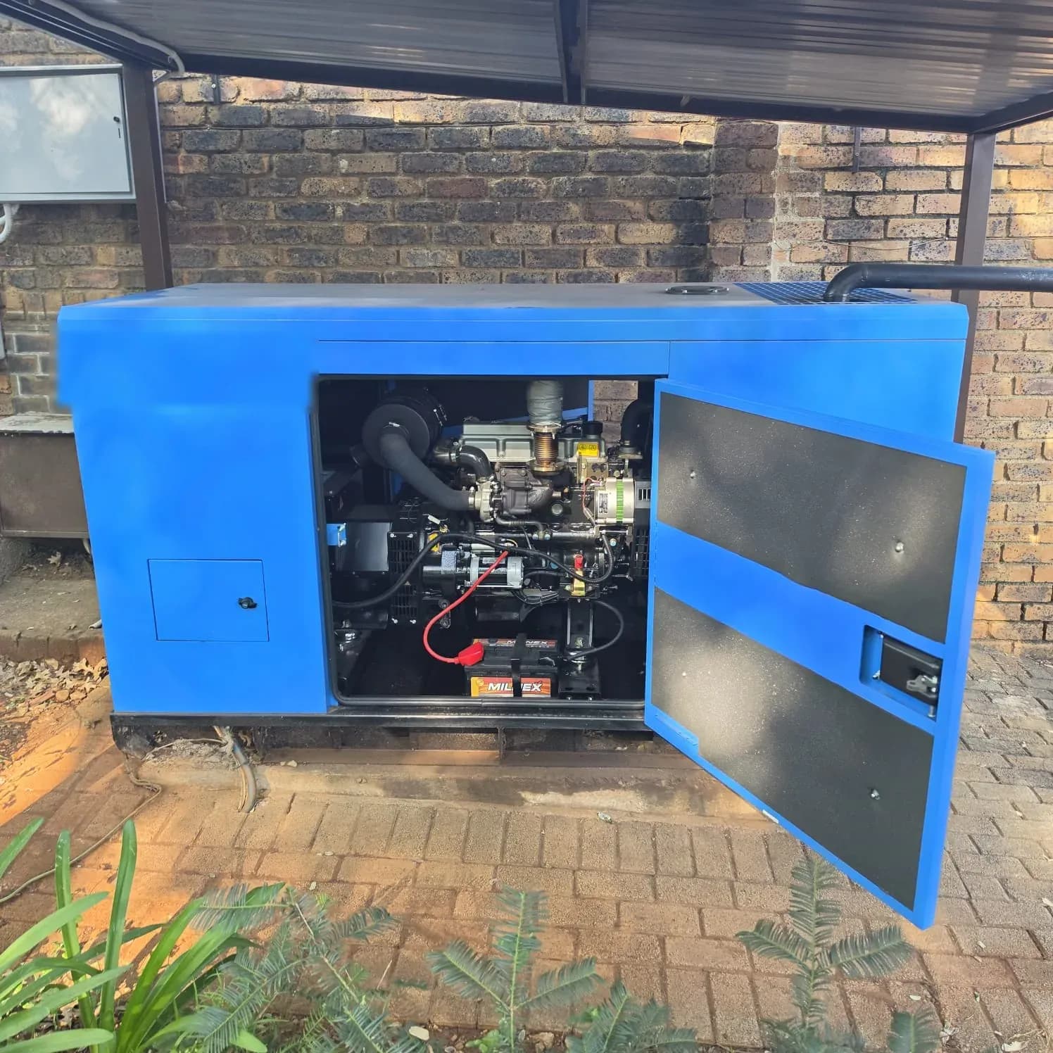 Blue Inyala Power canopied diesel generator with the door open on the engine and battery, under a carport