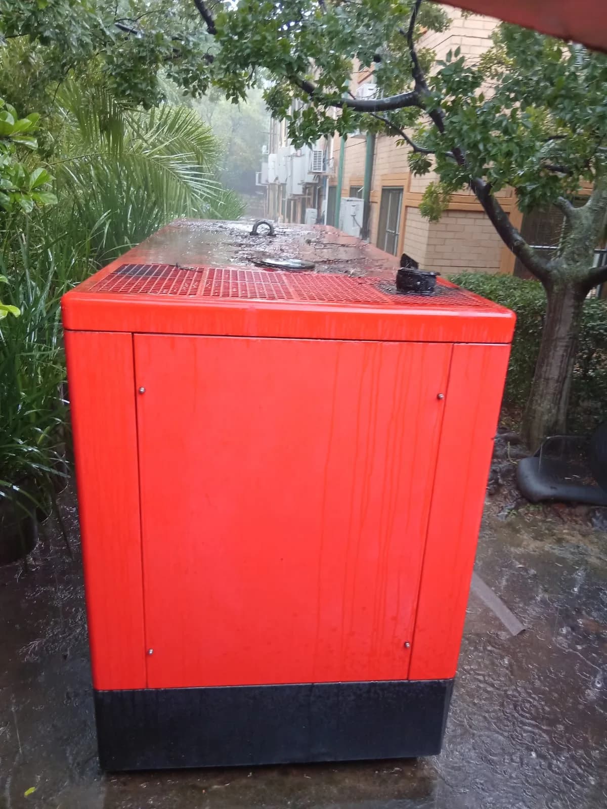 End of a red canopied diesel generator standing on wet paving in heavy rain under trees beside a block of units