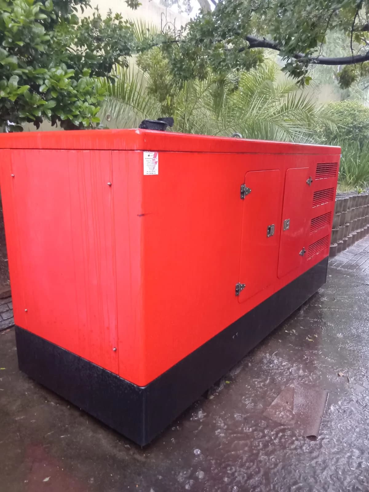 Side of a red canopied diesel generator with closed service doors on rain-soaked paving under trees