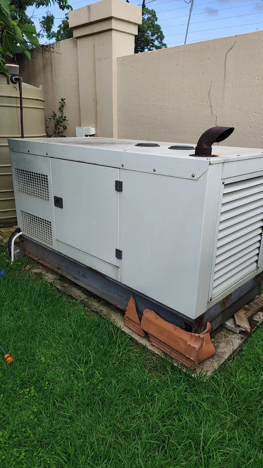 Cream canopied diesel generator raised on bricks on a lawn beside a boundary wall and water tank