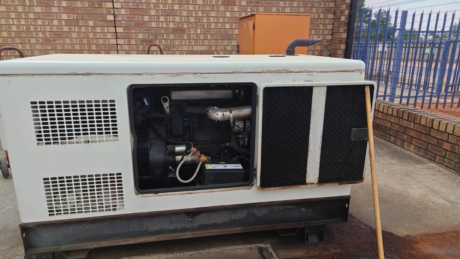White canopied diesel generator with a side door open on the engine and battery beside a face-brick wall