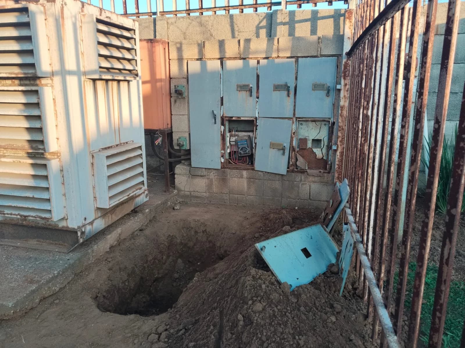Weathered louvred generator enclosure beside open electrical boxes, with a freshly dug cable trench in front