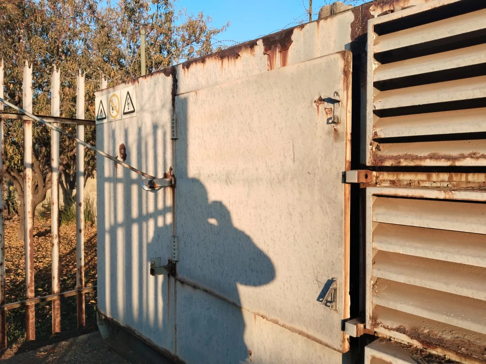 Rust-streaked louvred generator enclosure behind a palisade fence, the photographer's shadow on the door