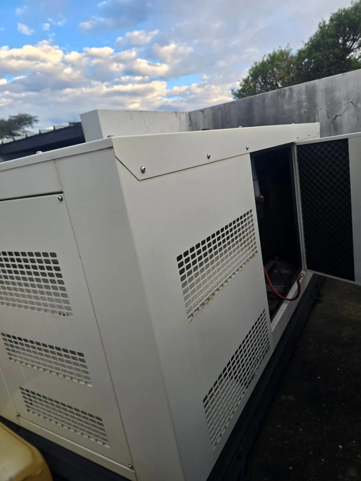 White canopied diesel generator with its end door open inside a walled enclosure under a cloudy sky