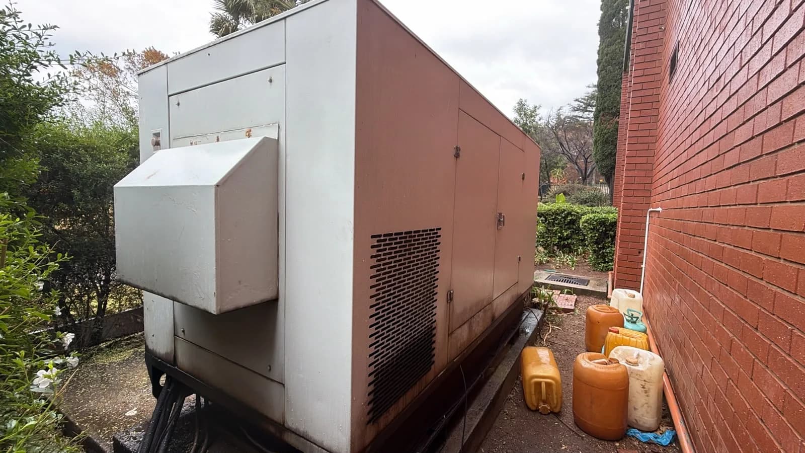 Large cream canopied diesel generator with an air outlet hood beside a red-brick wall and yellow fuel containers