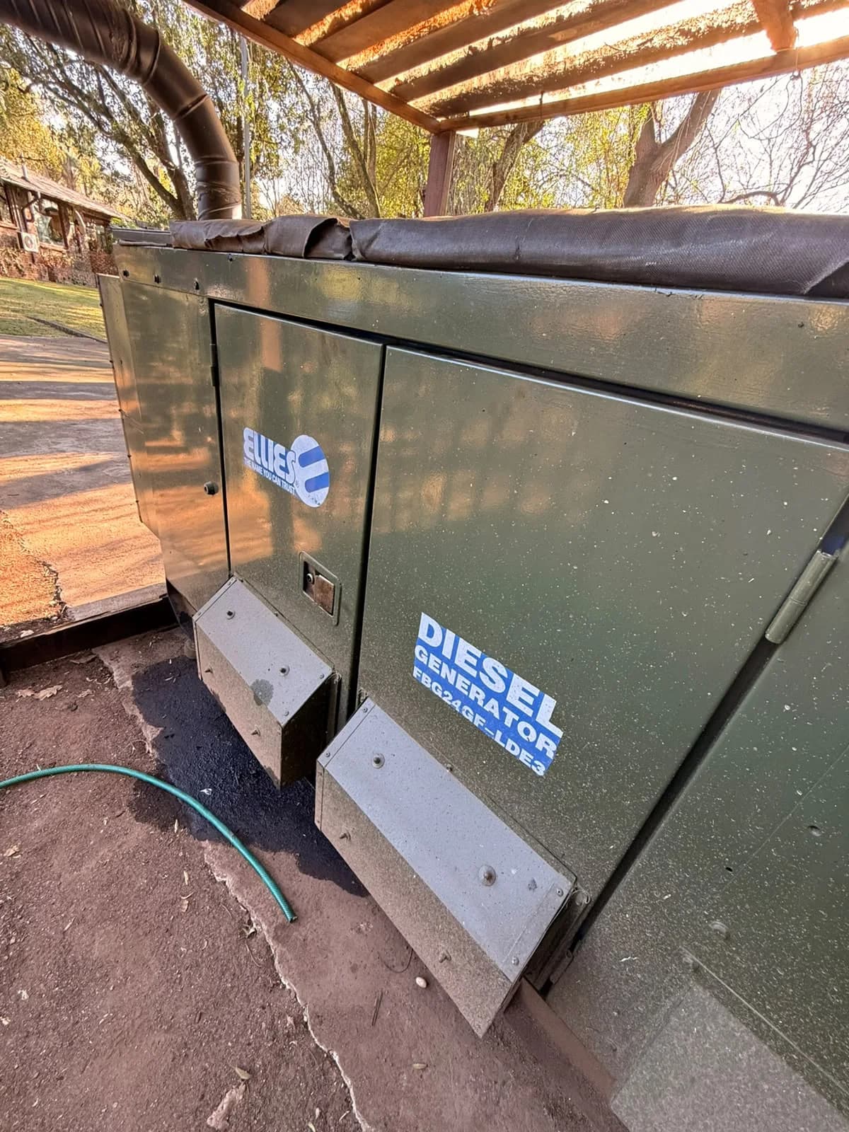 Dark green Ellies canopied diesel generator labelled FBG24GF-LDE3 under a timber pergola, a hose on the ground