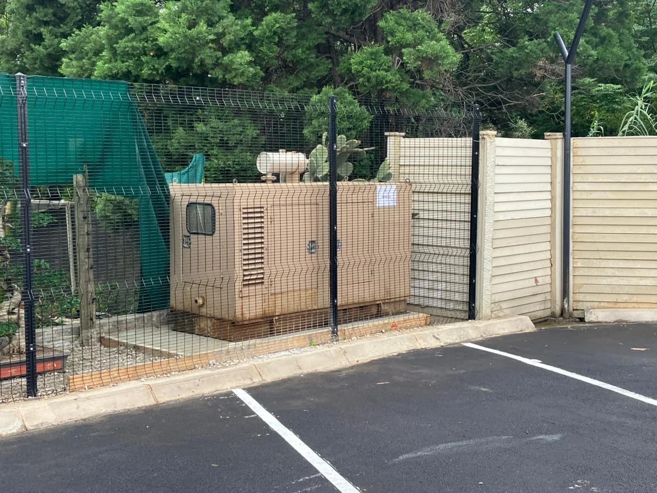 Beige canopied diesel generator inside a green mesh security fence beside a freshly tarred parking area