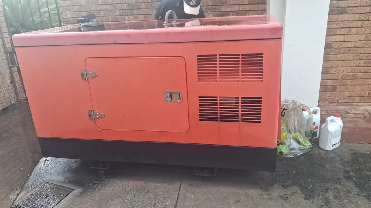 Faded red canopied diesel generator beside a face-brick wall with oil containers, one person behind it