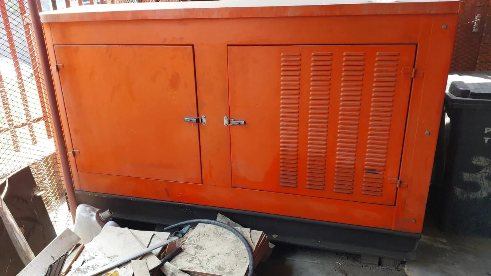 Orange canopied diesel generator with louvred doors beside a mesh fence, broken tiles and rubble in front