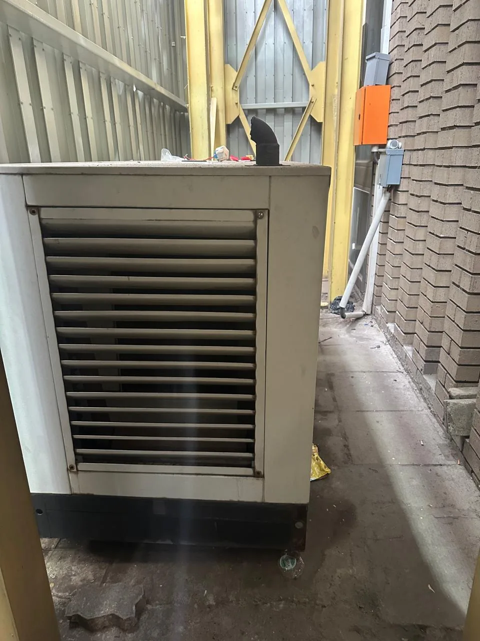 Louvred end of a cream canopied diesel generator in a narrow passage between a steel-clad building and brick wall
