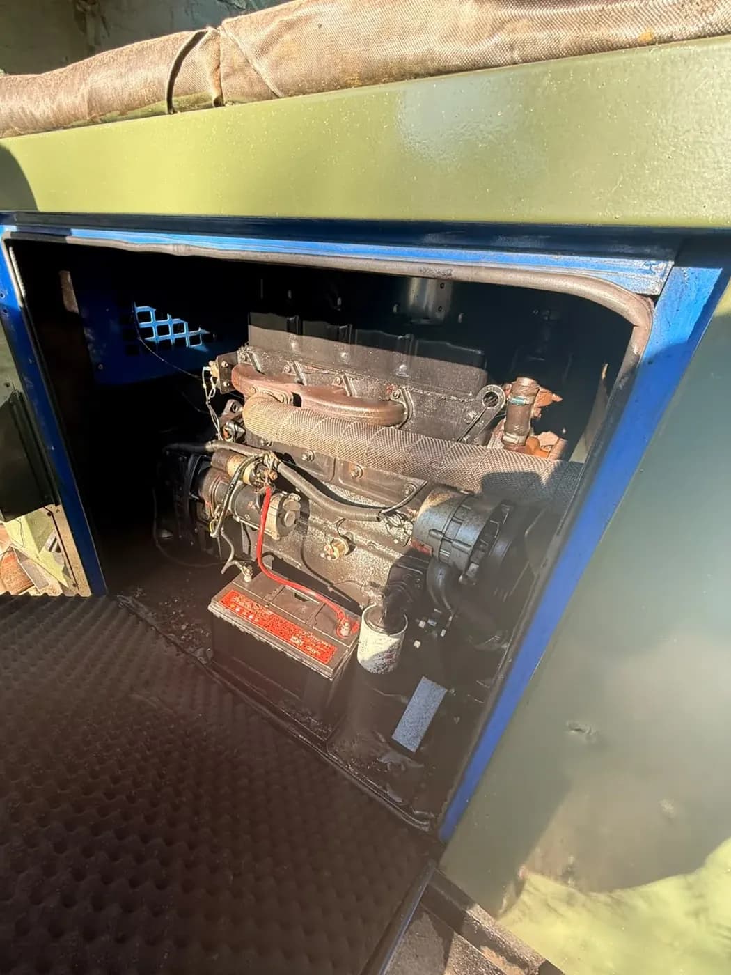 Olive-green canopy end door open on a small diesel engine and battery, with acoustic foam lining