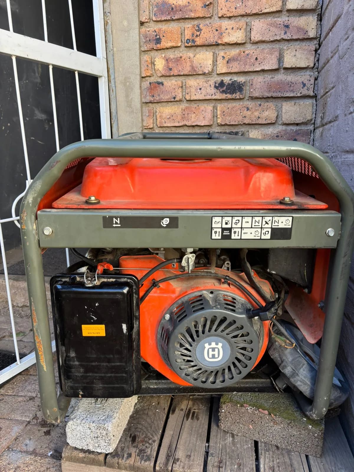 Orange-tanked portable generator in an olive tube frame, raised on bricks beside a face-brick wall
