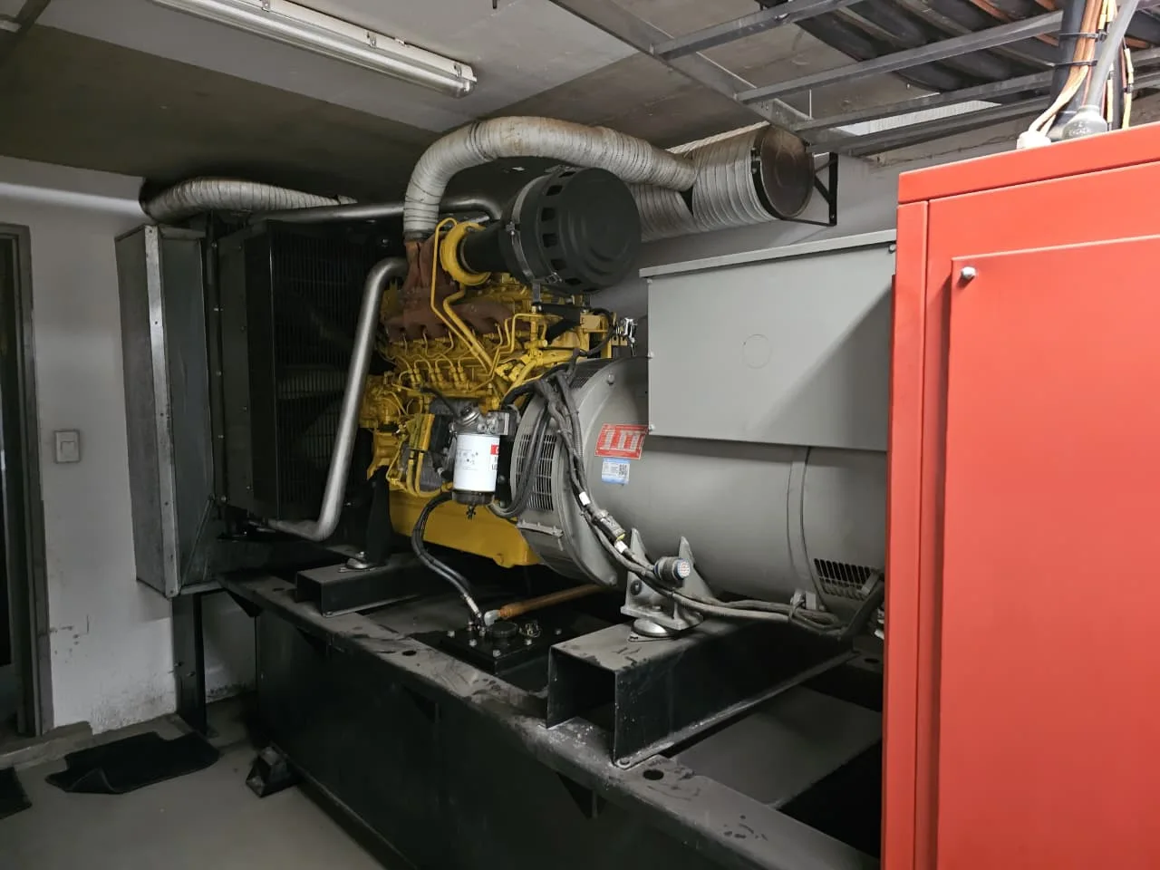 Yellow six-cylinder diesel engine and grey alternator on a skid in a plant room, red control cabinet at right