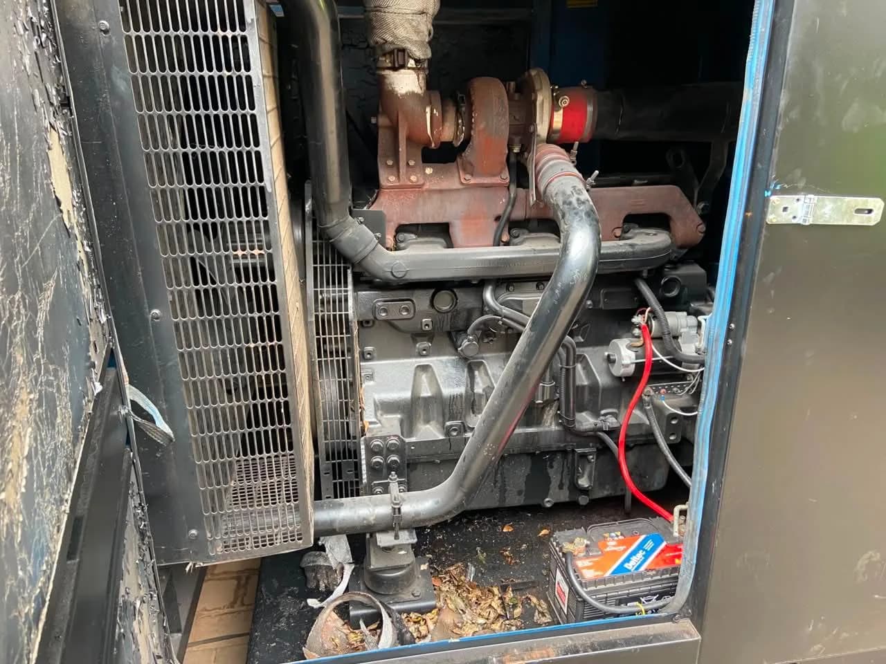 Six-cylinder turbo diesel engine inside an open canopy, radiator guard at left and a Deltec battery on the leafy floor