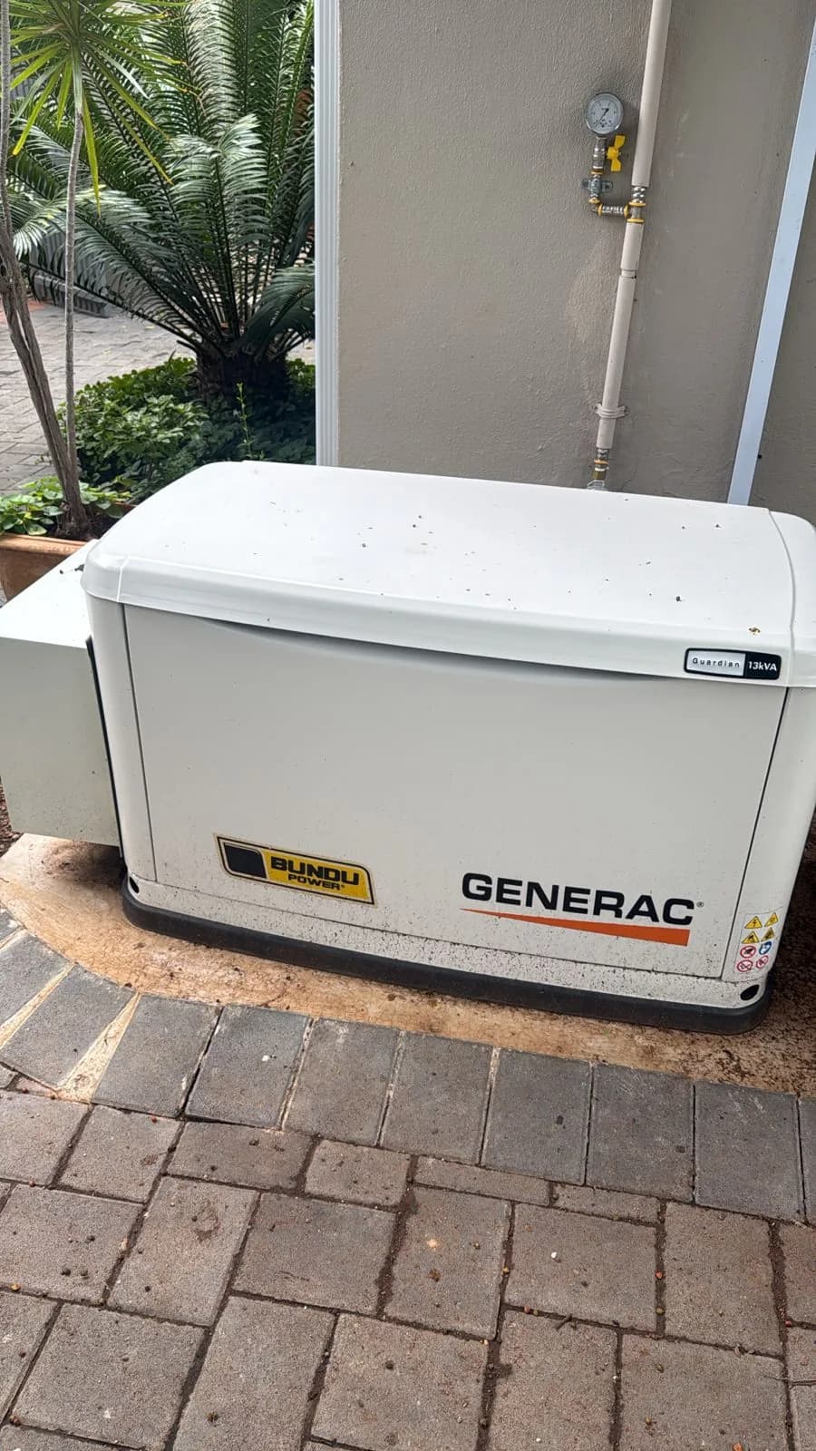 White Generac Guardian 13 kVA standby generator on a paved base beside a wall with a gas supply pipe and cycads