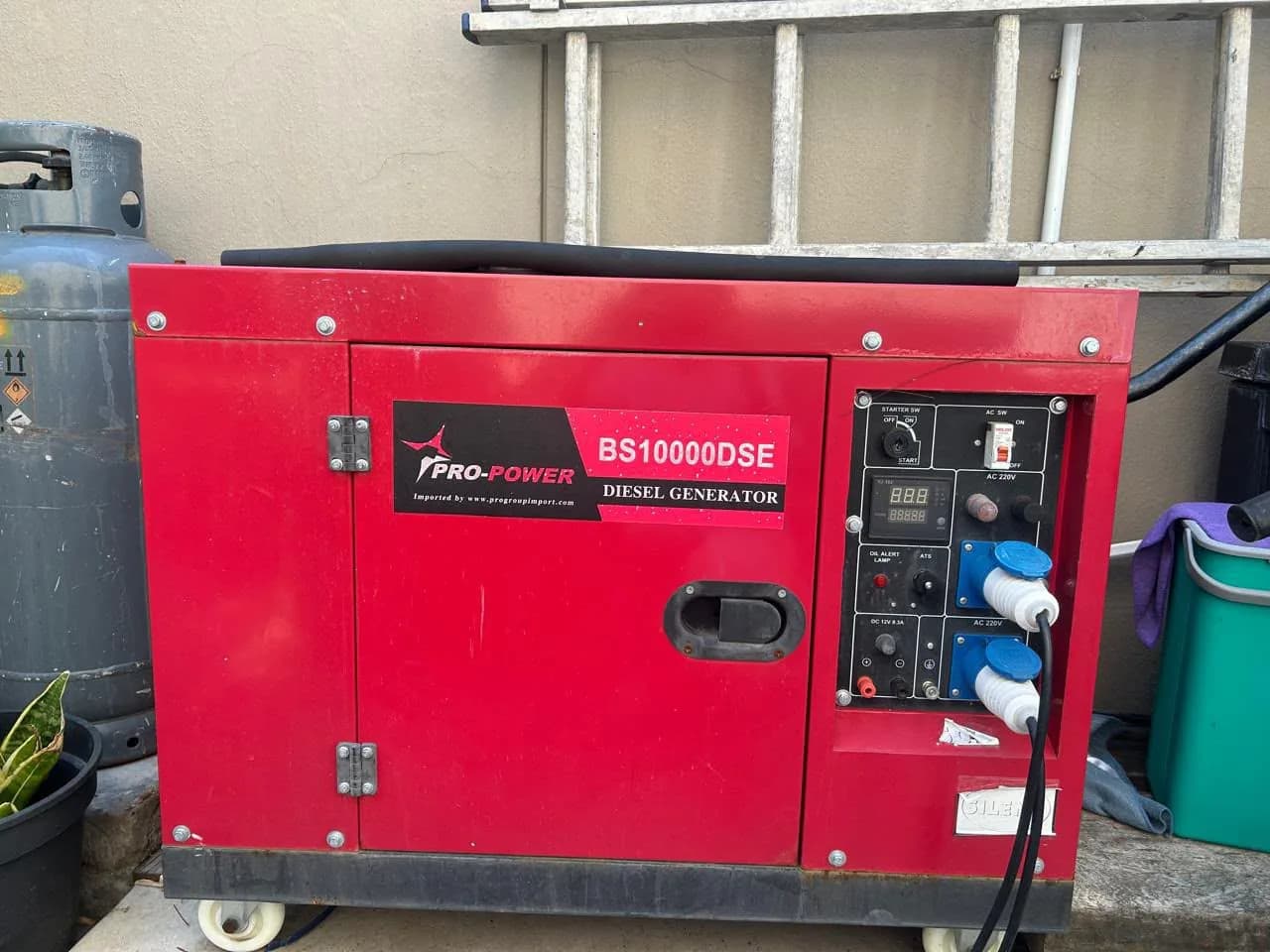 Red Pro-Power BS10000DSE silent diesel generator with two blue industrial sockets in use, beside a gas cylinder
