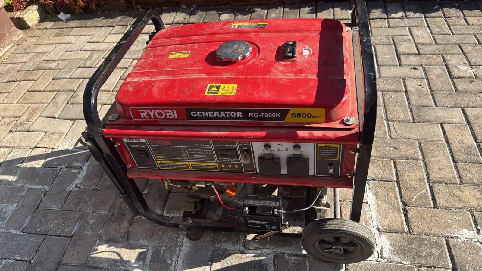 Red Ryobi RG-7900K 6800 W petrol generator with digital meter and sockets, on brick paving in sunlight