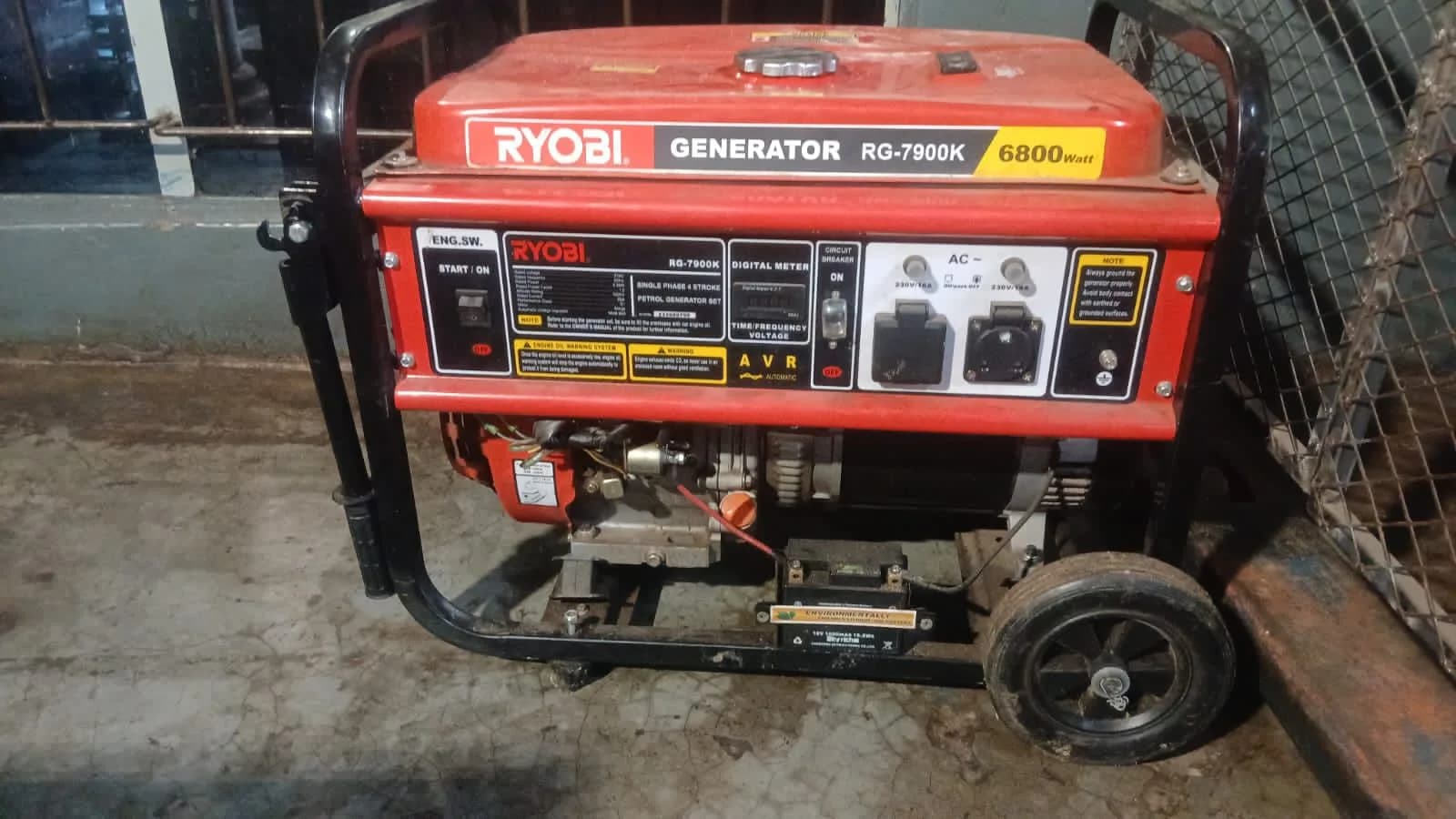 Red Ryobi RG-7900K 6800 W petrol generator showing its control panel, AVR and sockets beside a mesh fence
