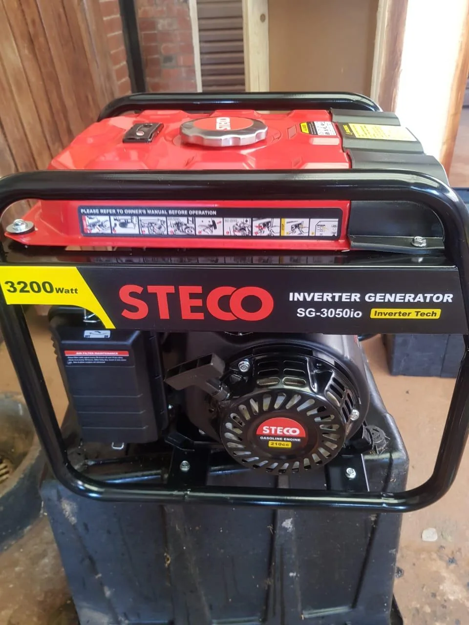 Red and black Steco SG-3050io 3200 W inverter generator with its recoil starter, standing on a black crate